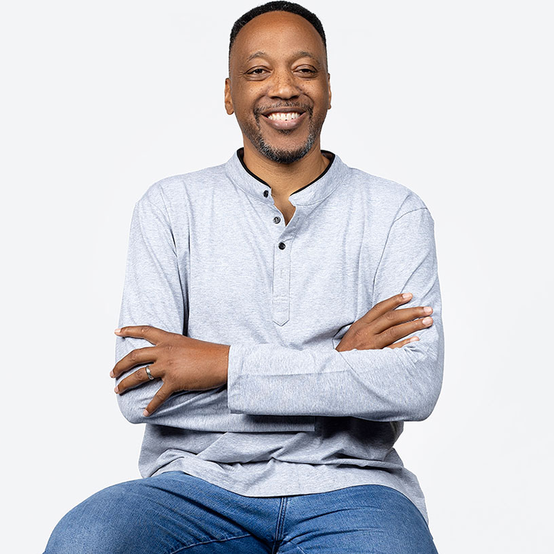 A smiling man in a gray shirt and jeans sitting on a chair.