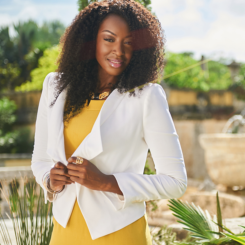 A black woman in a yellow dress and white blazer.