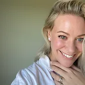 A person with blonde hair smiles, wearing a white top and a ring on their hand, set against a plain background.