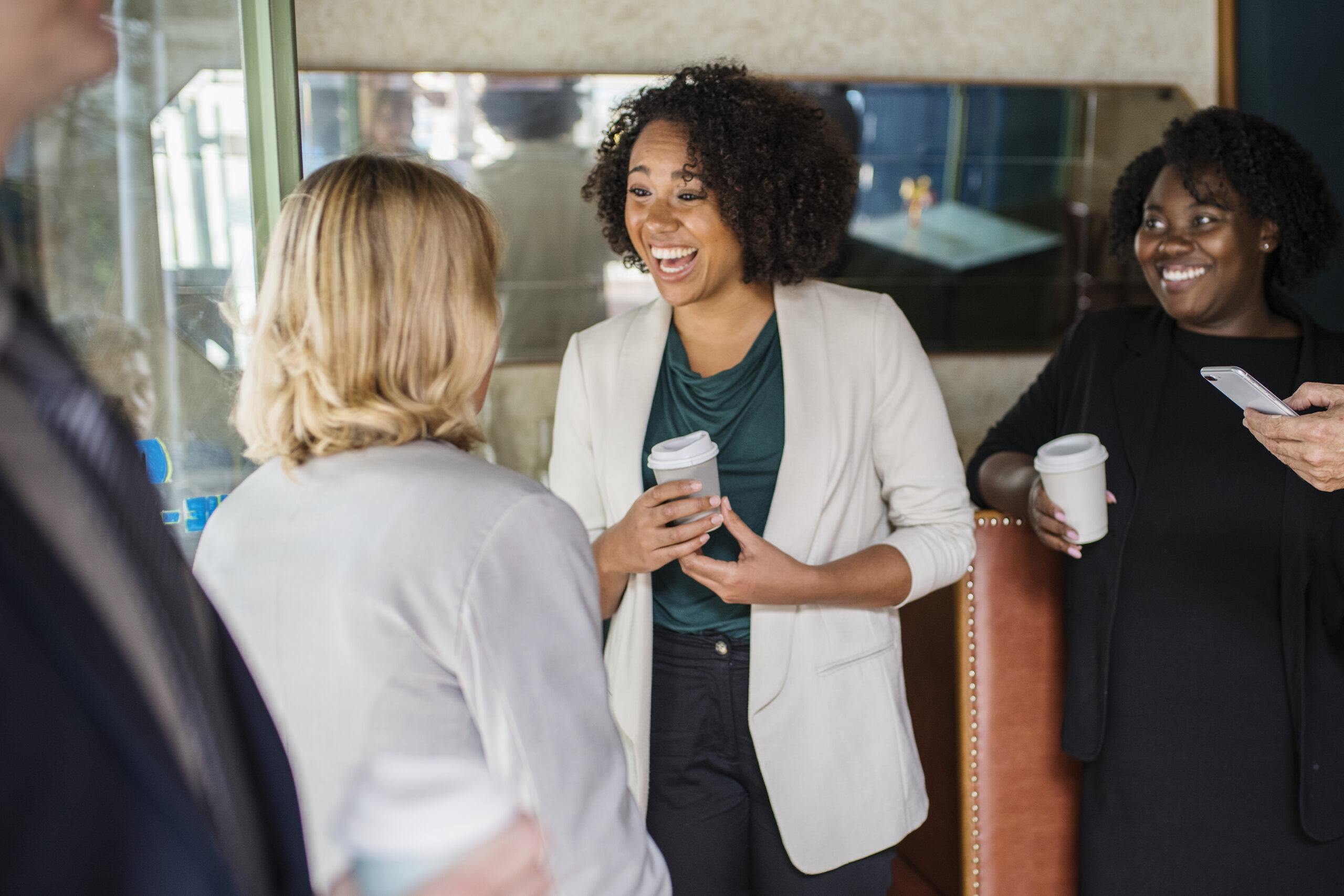 Four people in business attire stand together indoors, holding coffee cups and talking. Two women in the center are engaged in conversation, smiling.